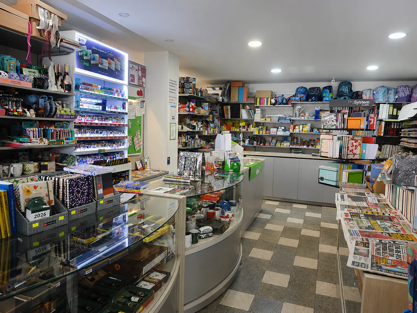 Interior amplo e organizado da Livraria e Papelaria Saimeiro em Oliveira do Bairro, destacando expositores com mochilas escolares, cadernos, jornais, canetas e material de escritório sobre o balcão.